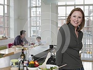 Woman Standing At Kitchen Counter With Family In Background