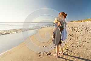 Woman is standing on a lonely beach with her child in her arms, looking into the sunset