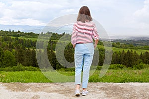 Woman standing in mountain, panoramic view of mountain range, trees. green grass. blue sky, female wearing casual trousers and