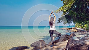 A woman standing on one leg while practicing yoga on wooden bridge over the sea during summer vacation.