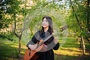 A woman is standing in the park among the trees with an acoustic guitar. Girl musician is resting and playing music in