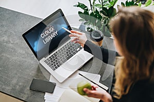 Woman is standing, pointing pencil on laptop computer with inscription on monitor- cloud library. Hipster girl working on pc