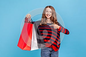 Woman standing pointing at shopping bags in her hands, looking at camera with happy expression.