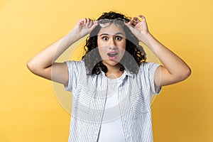 Woman standing raised eyeglasses and looking at camera with big eyes, sees something shocked.