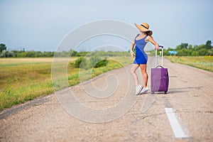 A woman is standing on the road with a hat and with a big bag. Girl in a blue tight-fitting dress with a purple suitcase