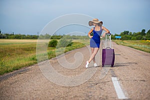 A woman is standing on the road with a hat and with a big bag. Girl in a blue tight-fitting dress with a purple suitcase