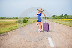 A woman is standing on the road with a hat and with a big bag. Girl in a blue tight-fitting dress with a purple suitcase