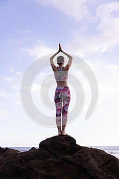 Woman standing on the rock, practicing yoga. Young woman raising arms with namaste mudra at the beach. Blue sky background. Yoga