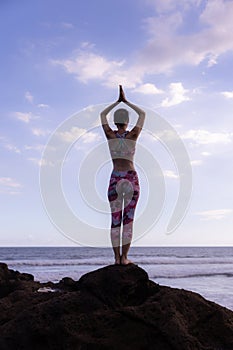 Woman standing on the rock, practicing yoga. Young woman raising arms with namaste mudra at the beach. Blue sky background. Yoga