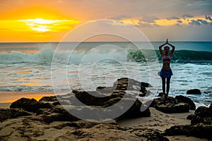 Woman standing on the rock, practicing yoga. Young woman raising arms witih namaste mudra at the beach. Bali