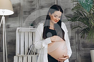 Woman standing and touching with hands her naked big belly.  on gray background. Emotional loving pregnancy time