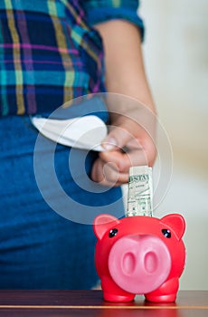 Woman standing up facing camera holding up empty denim pocket, pink piggy bank sitting on desk in front
