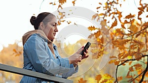 Woman stands on balcony against autumn park looking at phone