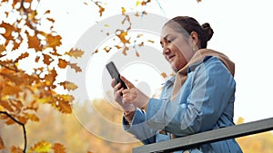 Woman stands on balcony against autumn park looking at phone