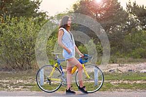 Woman stands with a bicycle on a road in the park in the sunset
