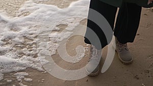 Woman stands in boots along sea foam blown by wind on sandy beach in slow motion. Female feet in waterproof footwear by