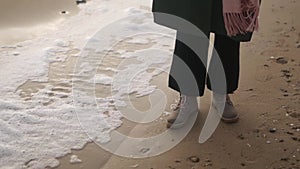 Woman stands in boots along sea foam blown by wind on sandy beach in slow motion. Female feet in waterproof footwear by