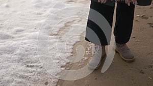 Woman stands in boots along sea foam blown by wind on sandy beach in slow motion. Female feet in waterproof footwear by