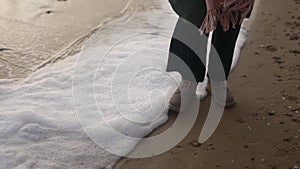 Woman stands in boots along sea foam blown by wind on sandy beach in slow motion. Female feet in waterproof footwear by