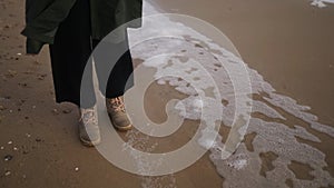 Woman stands in boots next to sea foam blown by wind on sandy beach in slow motion. Female feet in waterproof footwear