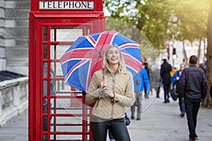 Woman stands in front of a red telephone booth with a British flag umbrella in London, United Kingdom