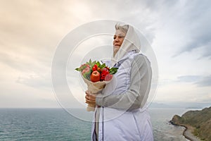 Woman stands on a high hill near the sea and looks into the distance with a bouquet in her hands