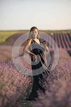 woman stands in a lavender field of purple flowers, holding a bouquet of flowers. The scene is serene and peaceful, with