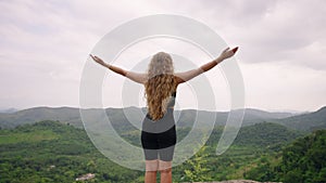 Woman stands on mountain peak, arms raised embracing vast green landscape. Solo traveler experiences wilderness