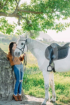 Woman stands near tree and holds white horse by the bridle