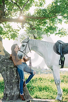 Woman stands near tree and holds white horse by the bridle