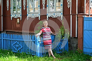 The woman stands near the wooden house with carved platbands