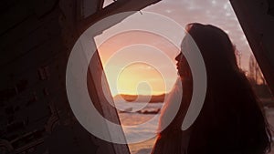 Woman stands in old wooden ruined leaning house interior on sea beach, looks at window on sunset. Mixed ethnic female