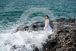 A woman stands on a rock in the sea during a storm. Dressed in a white long dress, the waves break on the rocks and