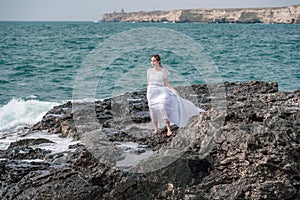 A woman stands on a rock in the sea during a storm. Dressed in a white long dress, the waves break on the rocks and