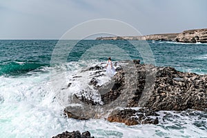 A woman stands on a rock in the sea during a storm. Dressed in a white long dress, the waves break on the rocks and