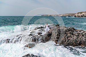 A woman stands on a rock in the sea during a storm. Dressed in a white long dress, the waves break on the rocks and