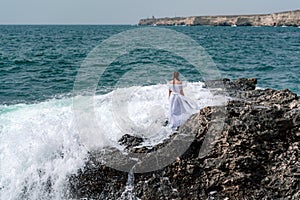 A woman stands on a rock in the sea during a storm. Dressed in a white long dress, the waves break on the rocks and