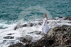 A woman stands on a rock in the sea during a storm. Dressed in a white long dress, the waves break on the rocks and