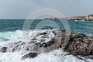 A woman stands on a rock in the sea during a storm. Dressed in a white long dress, the waves break on the rocks and
