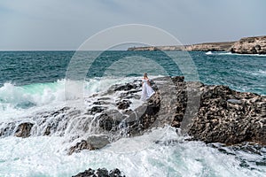 A woman stands on a rock in the sea during a storm. Dressed in a white long dress, the waves break on the rocks and