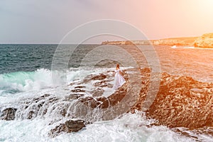 A woman stands on a rock in the sea during a storm. Dressed in a white long dress, the waves break on the rocks and