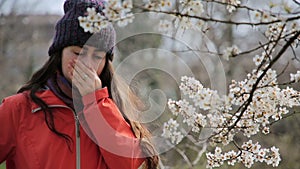 A woman stands on the street near a flowering tree, tries to smell the flowers on a branch, then sneezes and puts on a medical mas