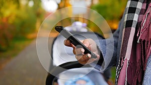 Woman stands with a stroller in the park autumn and dials a message on her phone. Maternity concept. Close up shot.
