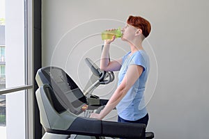 Woman stands on a treadmill in the gym and drinking water from a sports bottle