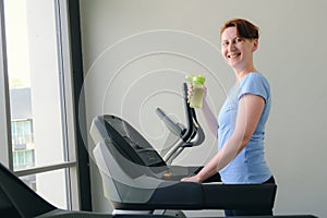 Woman stands on a treadmill in the gym, hold sports bottle with water and smiles