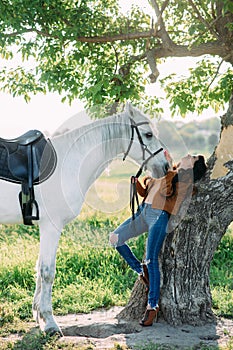 Woman stands in unbuttoned jacket near tree and holds white horse by the bridle