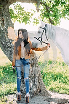 Woman stands in unbuttoned jacket near tree and holds white horse by the bridle