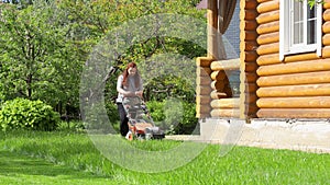 Woman starting lawn mower and begin mowing green lawn in backyard near wooden country house on summer sunny day