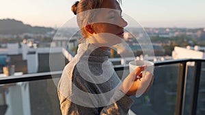 Woman starts her day with a cup of tea or coffee on the balcony at dawn. City landscape in the background