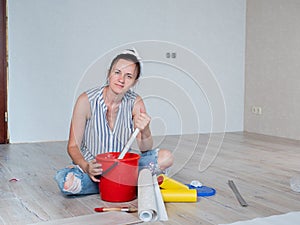 A woman stirs glue in a red bucket for wallpapering in an apartment.